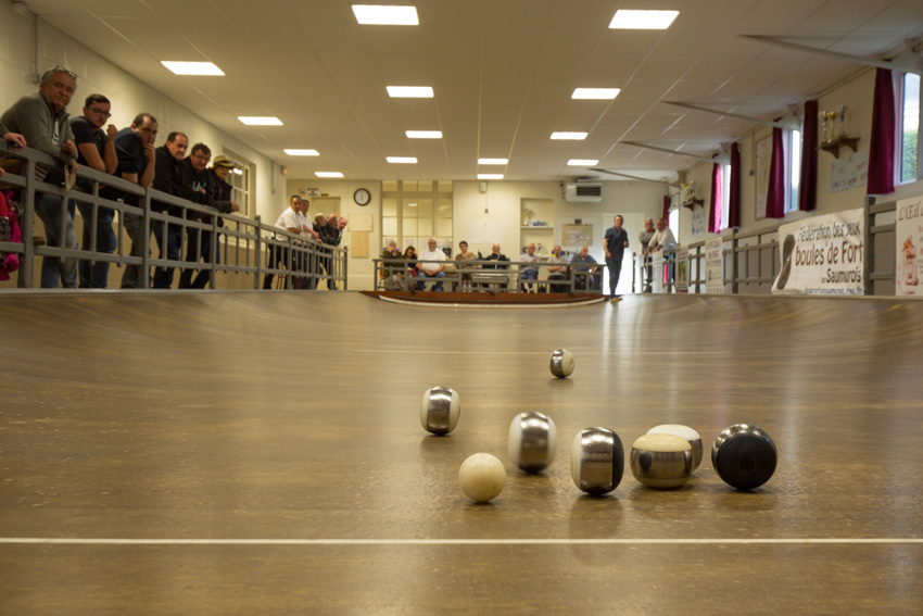 partie dans une société de boule de fort, un public assiste sur le côté gauche. en premier plan les boules roulent près du maitre. au fond, les joueurs